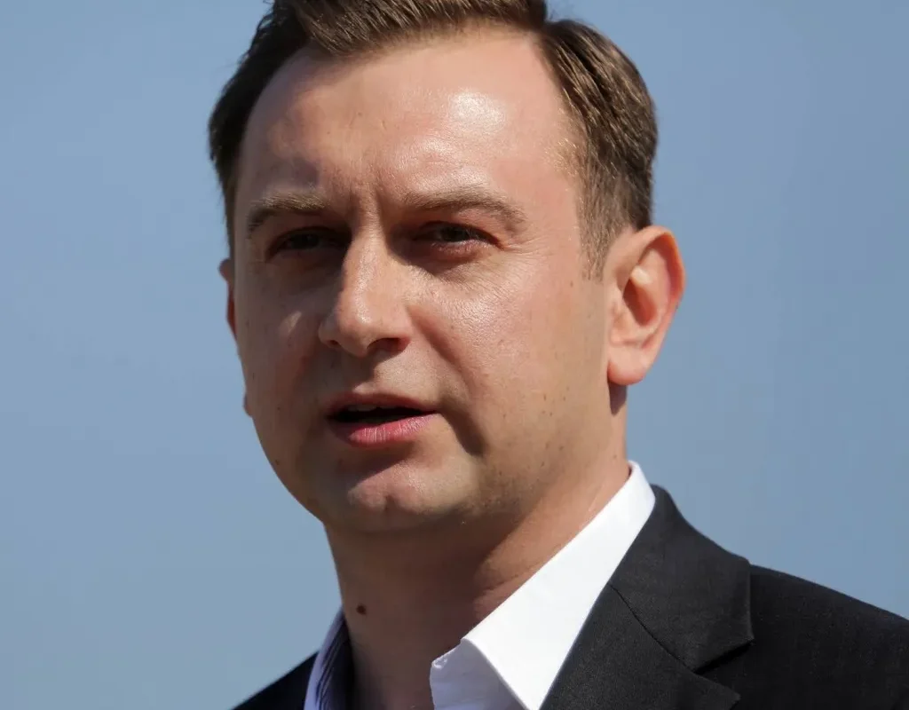 Zbliżenie na mężczyznę w marynarce i białej koszuli przemawiającego na zewnątrz na tle jasnego nieba.
