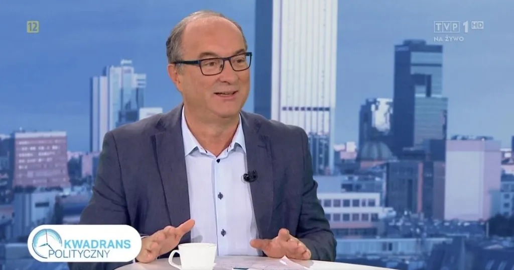 Mężczyzna w marynarce i okularach rozmawia w studiu telewizyjnym „Kwadrans Polityczny”, siedząc przy stole z filiżanką i dokumentami na tle panoramy miasta.