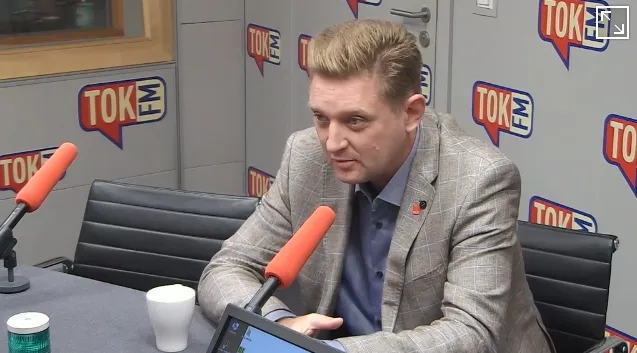 Mężczyzna w garniturze i niebieskiej koszuli siedzi w studiu radiowym TOK FM, rozmawiając przy dwóch pomarańczowych mikrofonach.