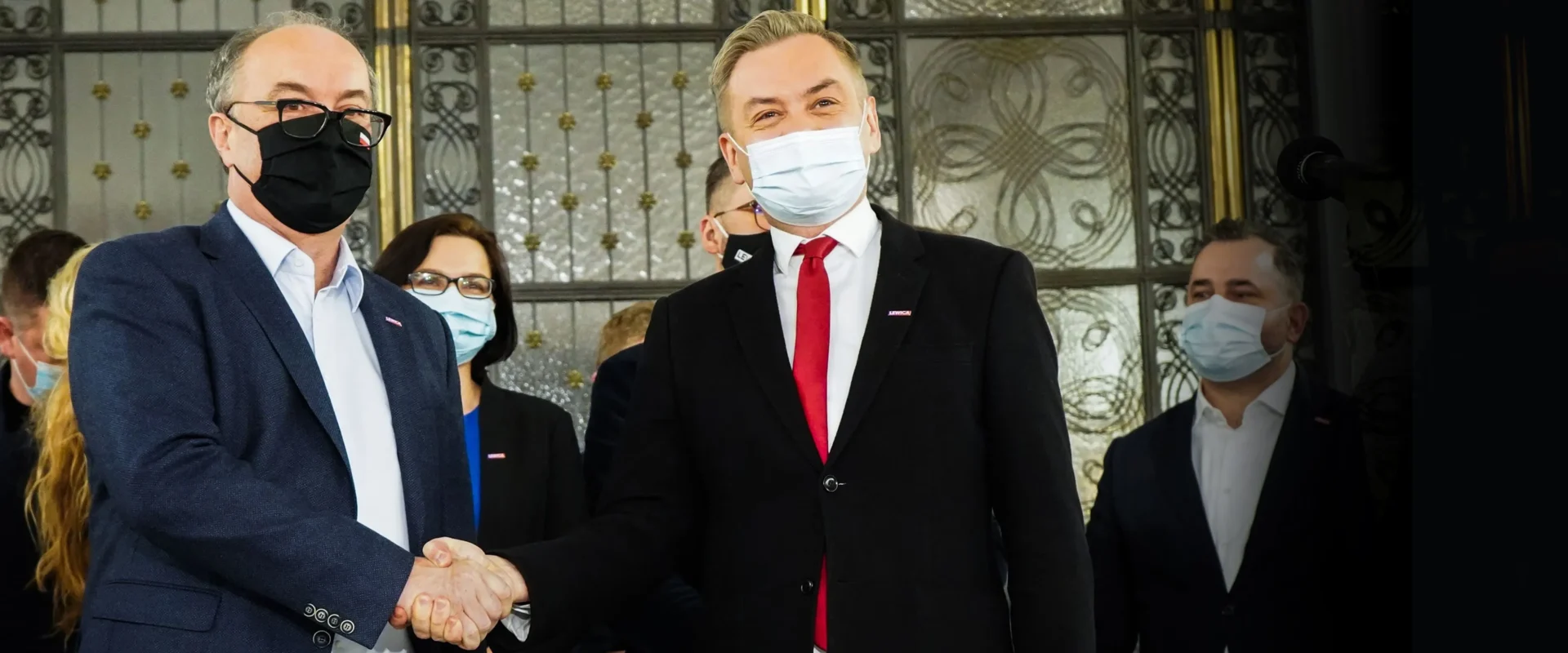 Dwoje mężczyzn w garniturach i maseczkach ściska sobie dłonie na tle ozdobnych drzwi, w tle stoją inni ludzie również w maseczkach.