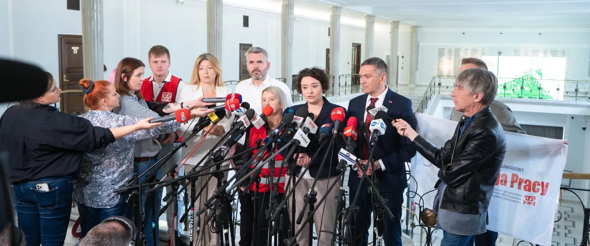Grupa osób w formalnych i casualowych strojach udziela konferencji prasowej, stojąc w otoczeniu licznych mikrofonów dziennikarzy w kontekście protestu „Nie dla handlu w niedzielę”.