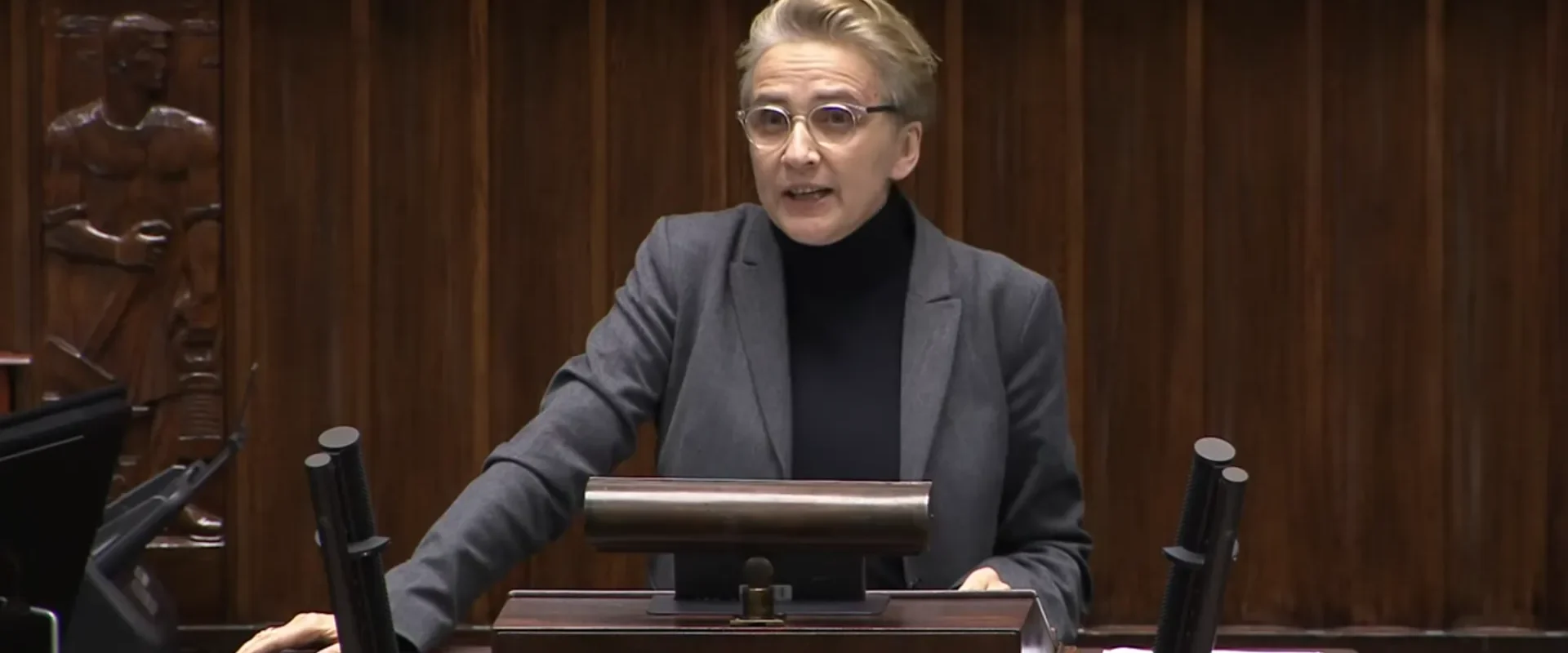 Kobieta w okularach, szarym garniturze i czarnym golfie stoi przy mównicy w sali plenarnej i przemawia, w tle drewniana dekoracja z mikrofonami.