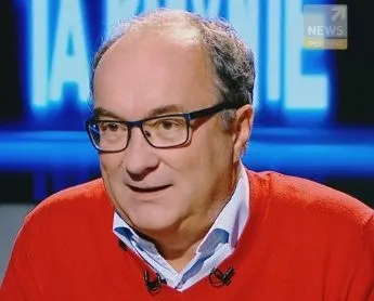 Mężczyzna w okularach i czerwonym swetrze z przypiętymi mikrofonami wypowiada się w studiu telewizyjnym.