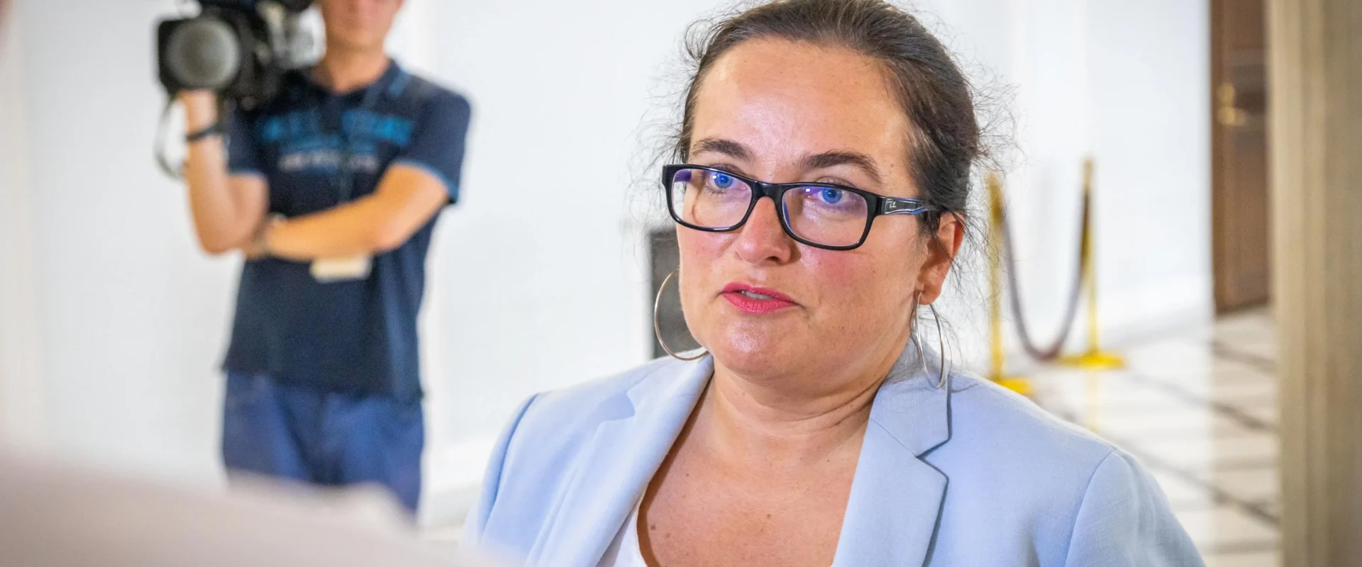 Kobieta w jasnoniebieskim żakiecie i czarnych okularach udziela wywiadu w holu; w tle stoi operator kamery.