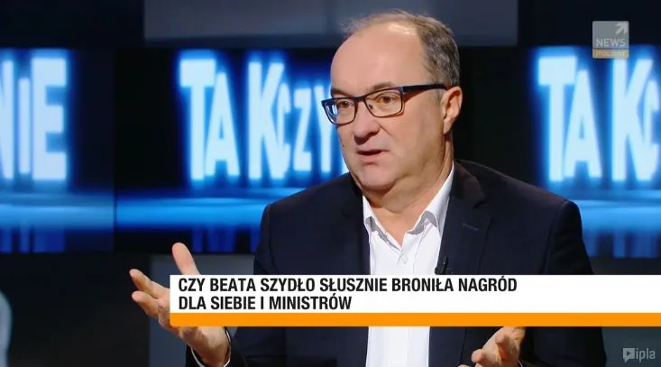 Mężczyzna w okularach i marynarce gestykuluje podczas telewizyjnego wywiadu, a na dole ekranu widoczny jest paskowy napis pytający, czy była premier słusznie broniła nagród dla siebie i ministrów.