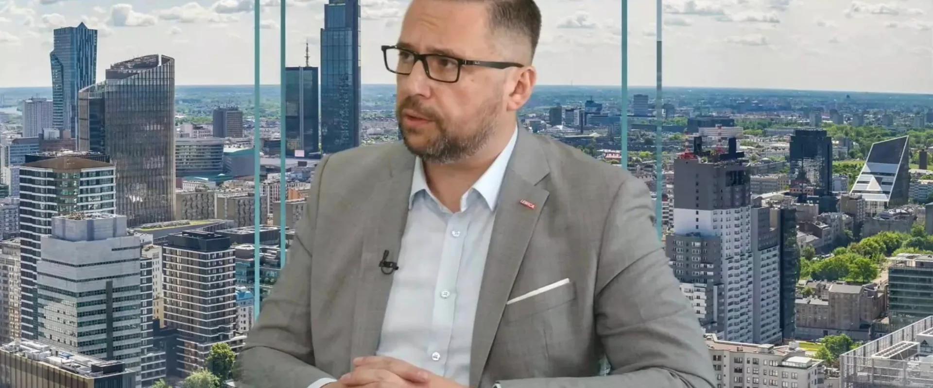 Mężczyzna w szarym garniturze i okularach siedzi przy stole w studiu telewizyjnym, a w tle widać panoramę miasta z wieżowcami.