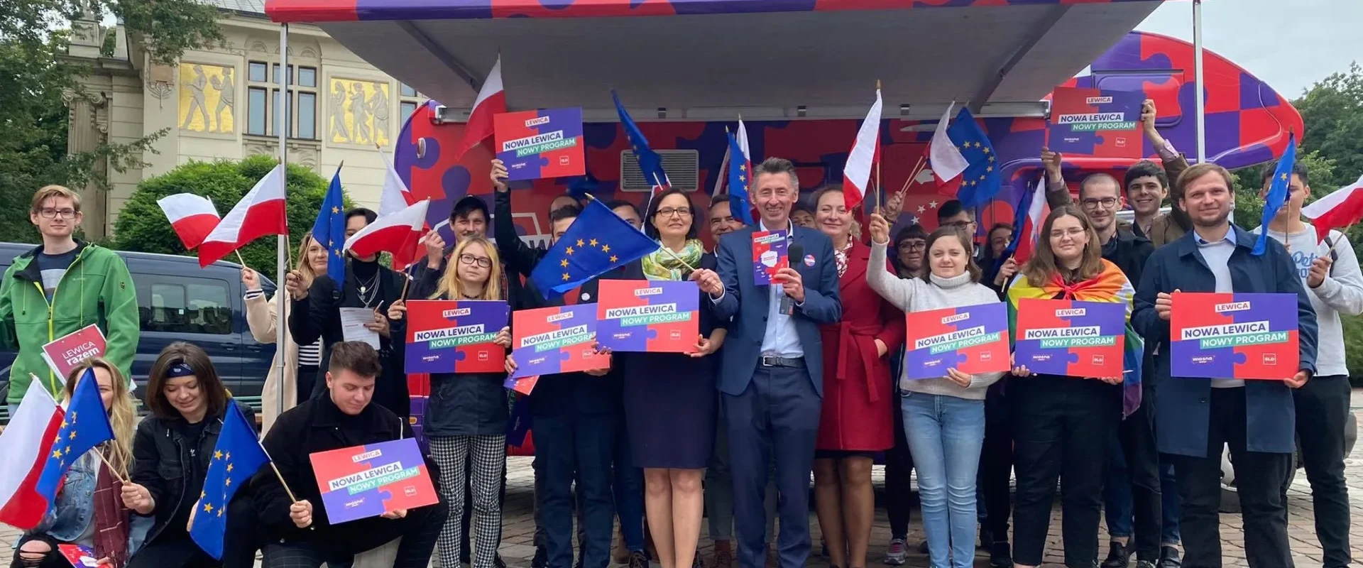 Grupa ludzi stoi przed kamperem kampanii Nowej Lewicy, trzyma plakaty „Nowa Lewica Nowy Program” oraz flagi Polski i Unii Europejskiej.
