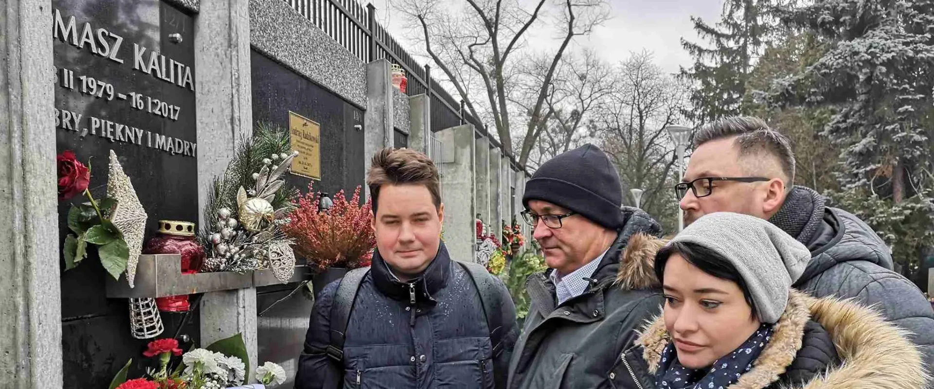 Grupa osób w zimowych kurtkach stoi przy kamiennym pomniku z tablicą upamiętniającą zmarłą osobę i składa kwiaty.