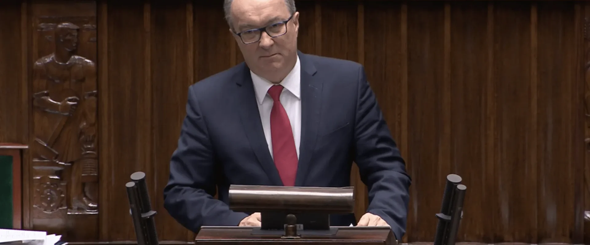 Mężczyzna w garniturze z czerwonym krawatem przemawia z mównicy w tle drewnianych paneli i rzeźb.