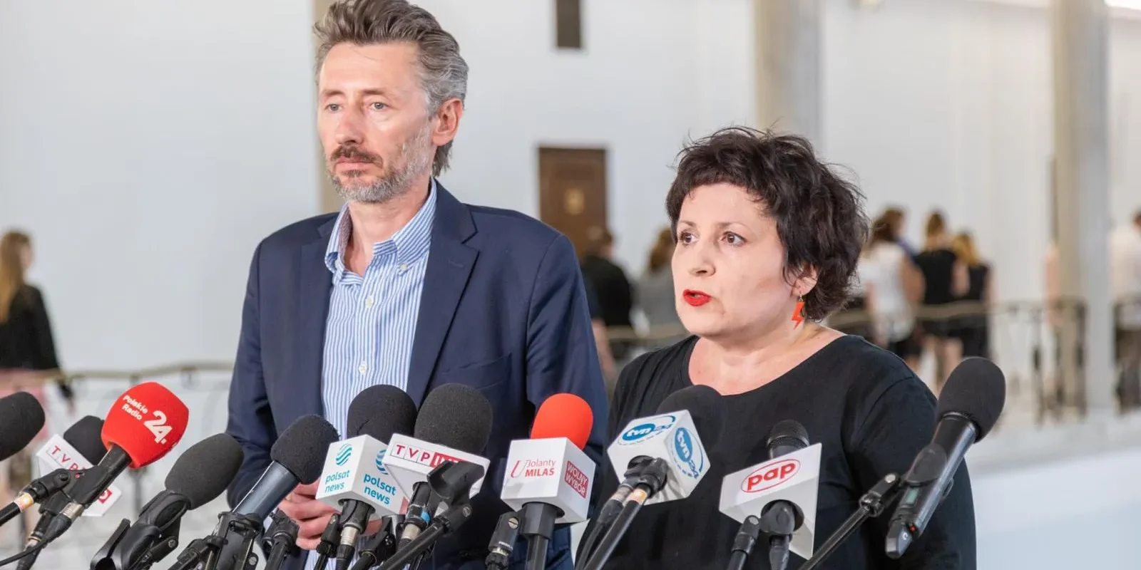 Dwoje dorosłych stoi przy licznym zestawie mikrofonów podczas konferencji prasowej w jasnym wnętrzu; kobieta przemawia, obok niej stoi mężczyzna.