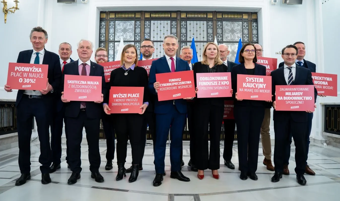Grupa polityków Lewicy w eleganckiej sali trzyma czerwone plakaty z hasłami o podwyżkach płac i polityce socjalnej, na tle flag, podczas rocznicowej okazji.