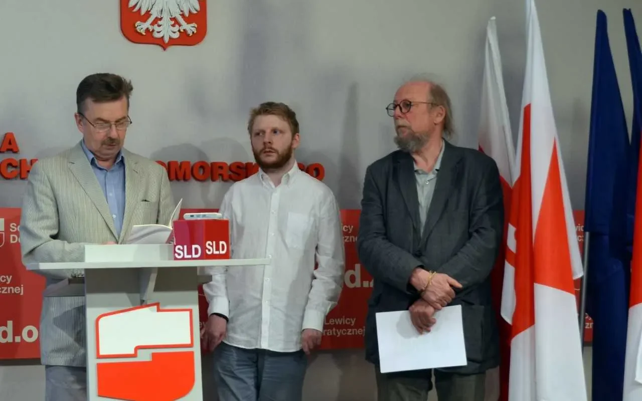 Trzech mężczyzn stoi przy podium z czerwonym logo SLD; jeden czyta z dokumentów, a w tle widoczne są flagi i godło Polski na ścianie.