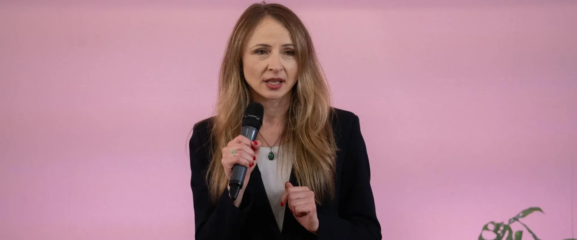 Kobieta w czarnym żakiecie trzyma mikrofon i przemawia podczas konferencji na tle różowej ściany, omawiając projekt prawa o zabójstwie drogowym.