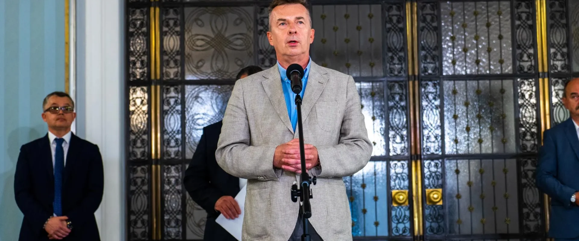 Mężczyzna w jasnoszarym żakiecie przemawia do mikrofonu podczas konferencji o zwiększeniu środków na badania i rozwój, w tle stoją inni mężczyźni w garniturach i ozdobne drzwi z żelaznymi ornamentami.