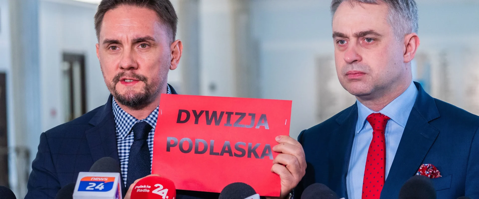 Na zdjęciu dwóch mężczyzn stoi przed mikrofonami, trzymając czerwony plakat z napisem "DYWIZJA PODLASKA".