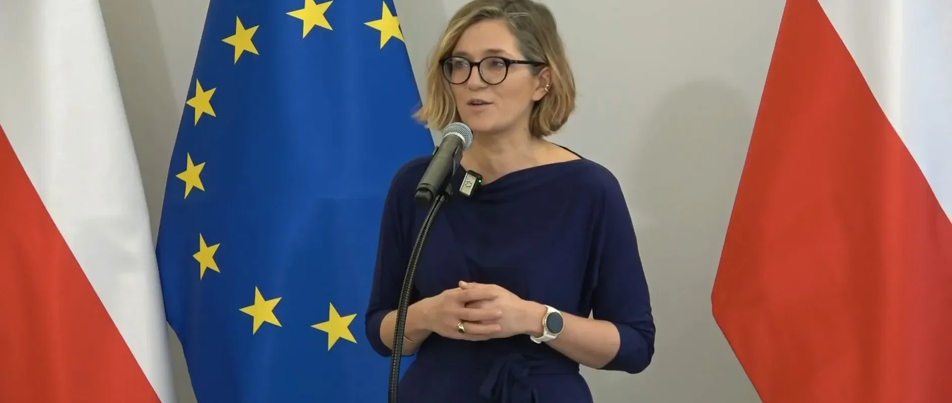 Kobieta w okularach przemawia do mikrofonu podczas konferencji, stojąc między flagą Unii Europejskiej a flagą Polski.