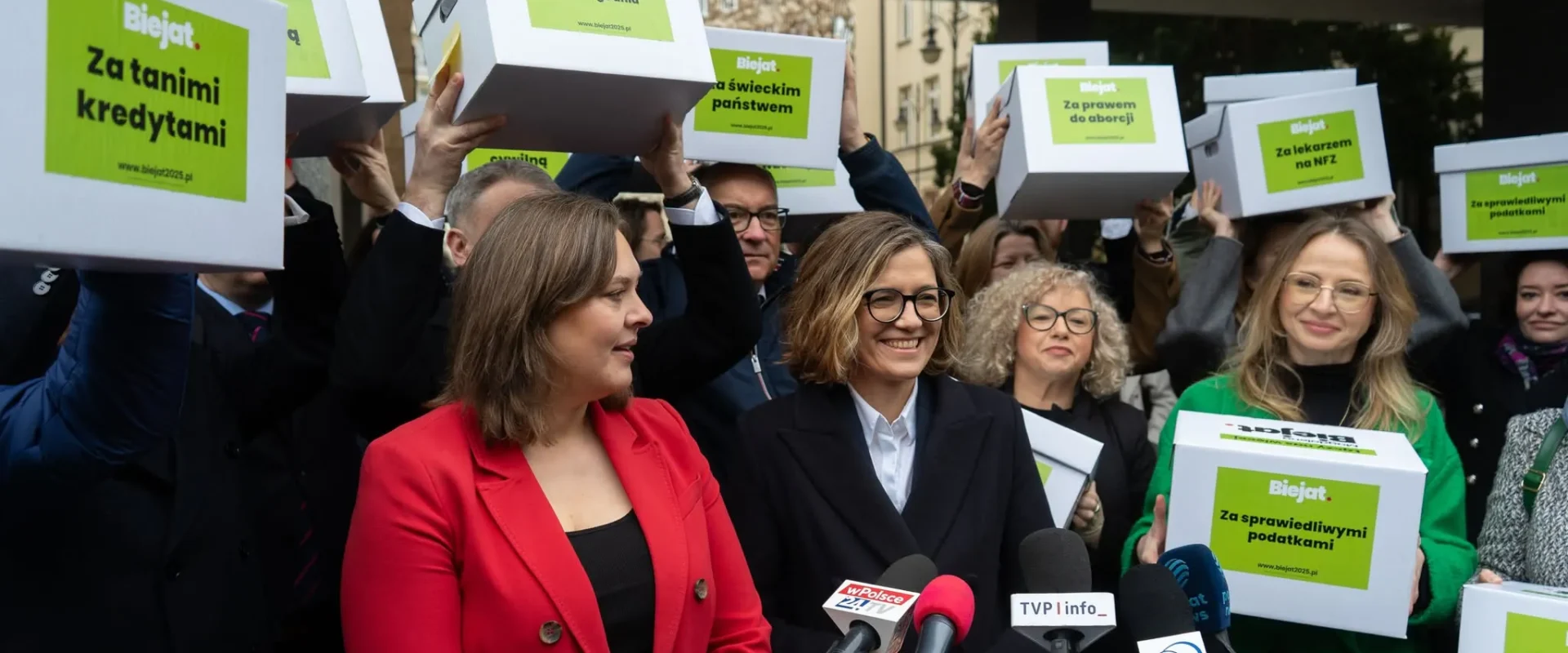 Grupa osób na konferencji prasowej trzyma nad głowami pudełka z zielonymi etykietami kampanii Magdaleny Biejat, na pierwszym planie kobieta w czerwonej marynarce oraz zestaw mikrofonów.