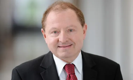 Portret mężczyzny w średnim wieku w ciemnym garniturze, białej koszuli i czerwonym krawacie, uśmiechnięty na jasnym, rozmytym tle.