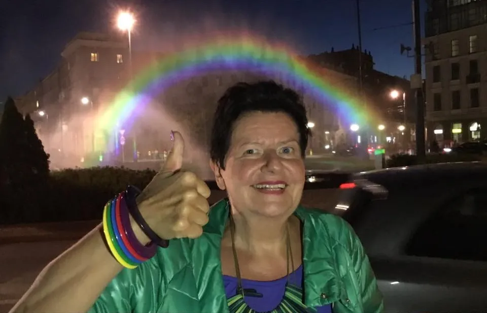 Uśmiechnięta kobieta w zielonej kurtce z kolorowymi bransoletkami na nadgarstkach na tle nocnego miasta i tęczowego łuku z fontanny.