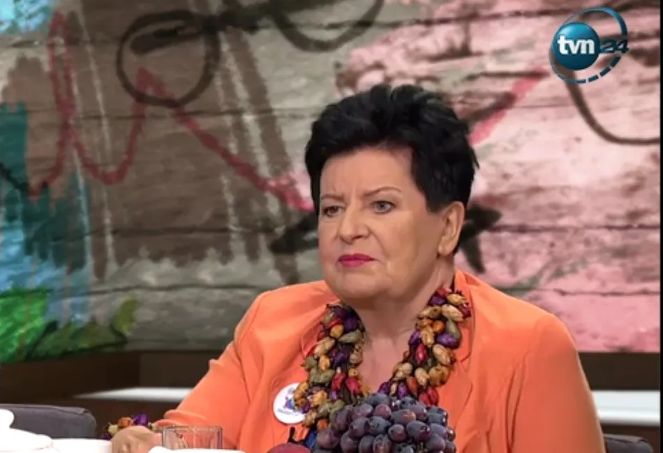 Kobieta z krótkimi czarnymi włosami w pomarańczowej marynarce i dużym naszyjnikiem z orzechów siedzi w studiu telewizyjnym, na tle graffiti i logotypu TVN24.