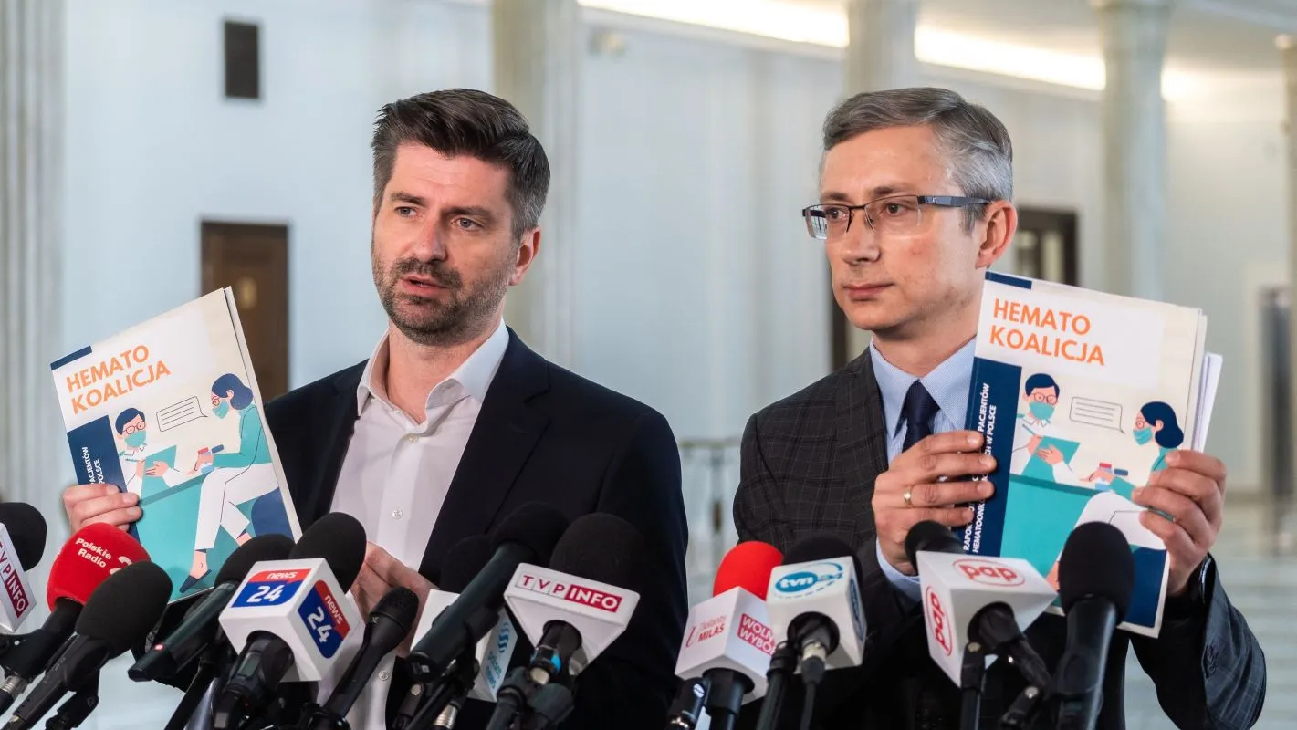 Dwóch mężczyzn w garniturach podczas konferencji prasowej trzyma ulotki z napisem „HEMATO KOALICJA” i stoi wśród mikrofonów.