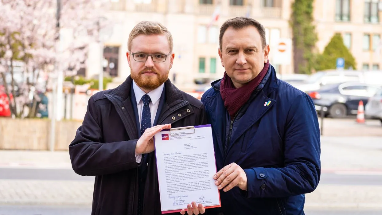 Dwóch mężczyzn trzymając wspólnie dokument, stoi na ulicy w tle z kwitnącymi drzewami.