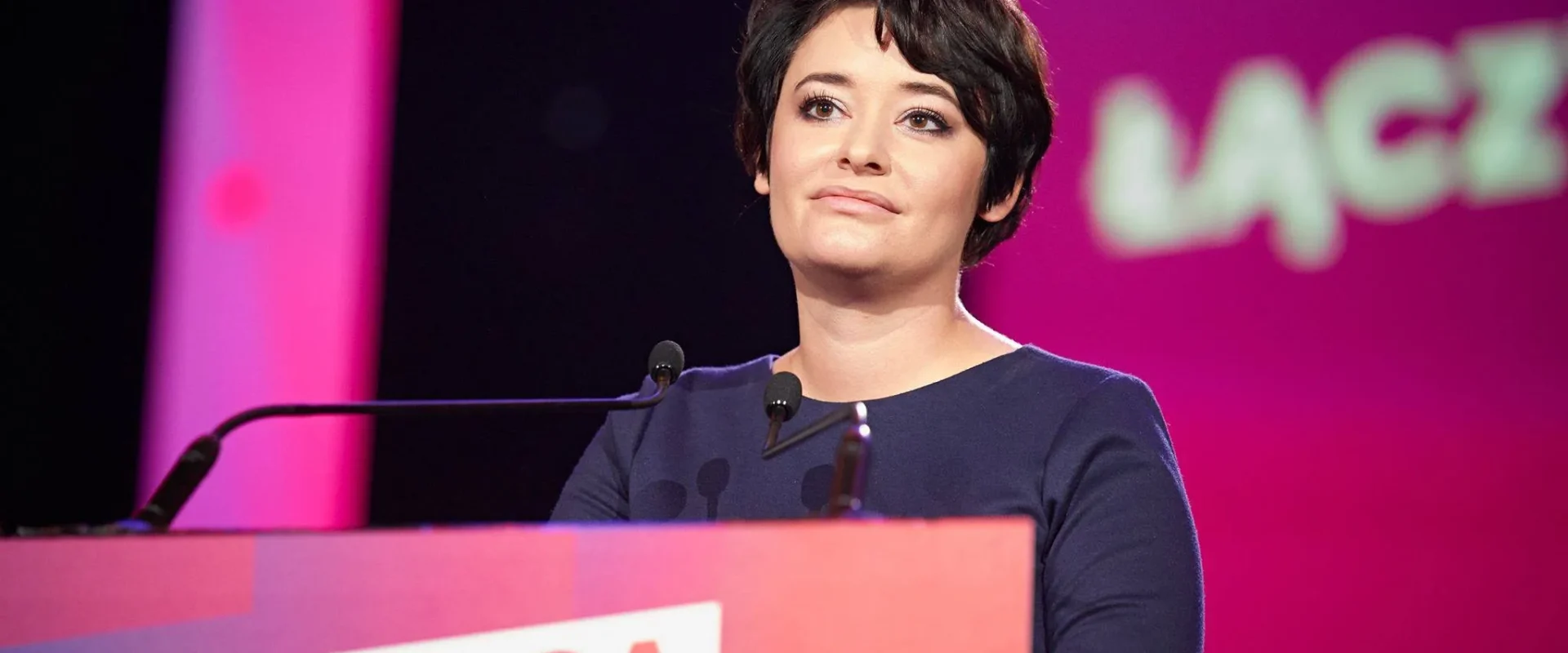 Kobieta stoi za podiumem i przemawia na scenie podczas wydarzenia Lewicy, w tle różowo-fioletowe oświetlenie oraz baner z napisem LEWICA.