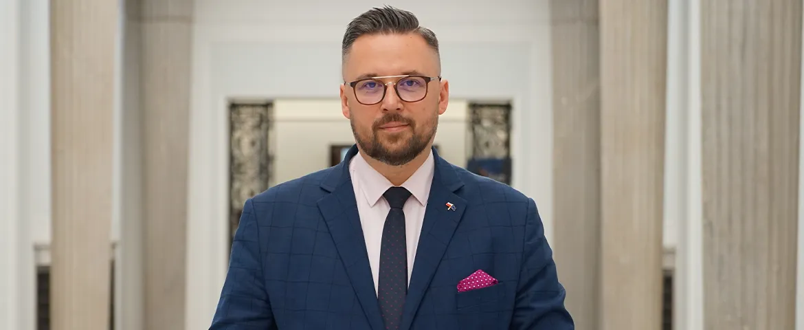 Mężczyzna w granatowym garniturze, różowej koszuli i różowej poszetce stoi w holu z kolumnami.