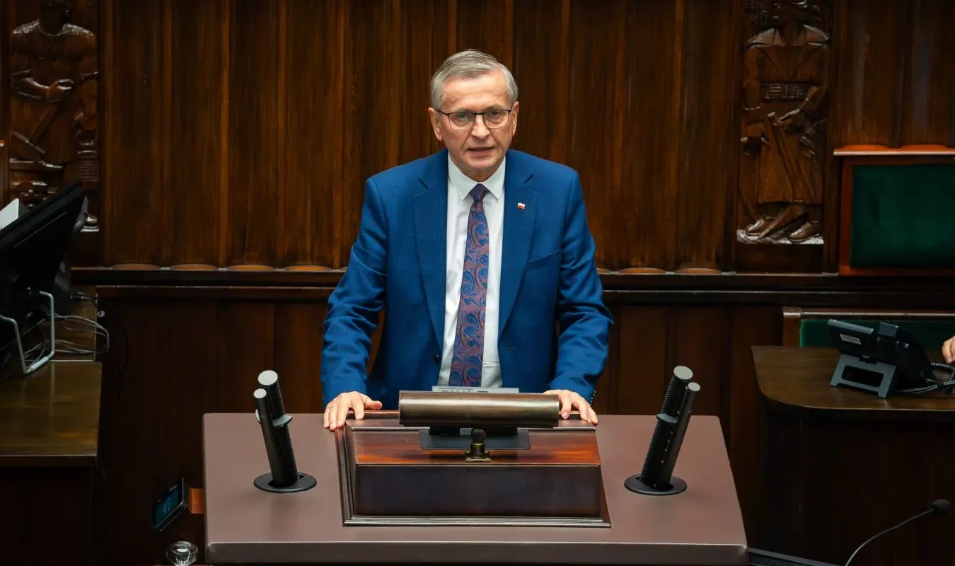 Polityk w niebieskim garniturze stoi przy mównicy w sali obrad podczas debaty budżetowej dotyczącej dodatkowych 55 mld zł na ochronę zdrowia przez dwa lata.