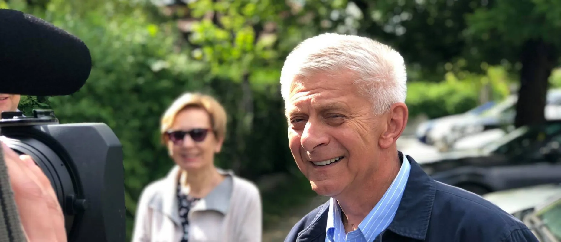 Mężczyzna o siwych włosach uśmiecha się na zewnątrz podczas nagrania; z lewej strony widoczna kamera filmowa, w tle osoba w okularach przeciwsłonecznych i zielone rośliny.