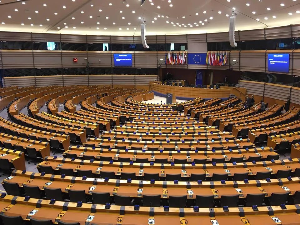 Widok sali plenarnej Parlamentu Europejskiego w Brukseli — rzędy drewnianych ław ustawionych w półokrąg, pulpit na środku i flagi Unii Europejskiej w tle.
