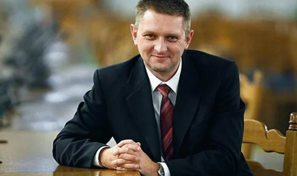 Mężczyzna w czarnym garniturze i czerwonym krawacie siedzi przy stole, złożone dłonie na blacie i uśmiecha się do kamery.