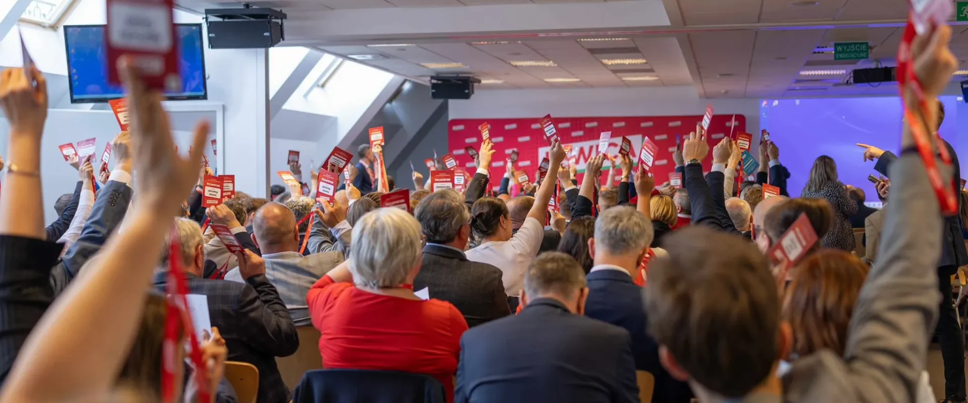 Sala konferencyjna pełna uczestników Kongresu Nowej Lewicy, którzy podnoszą czerwone kartki z poparciem, na tle sceny i banerów.