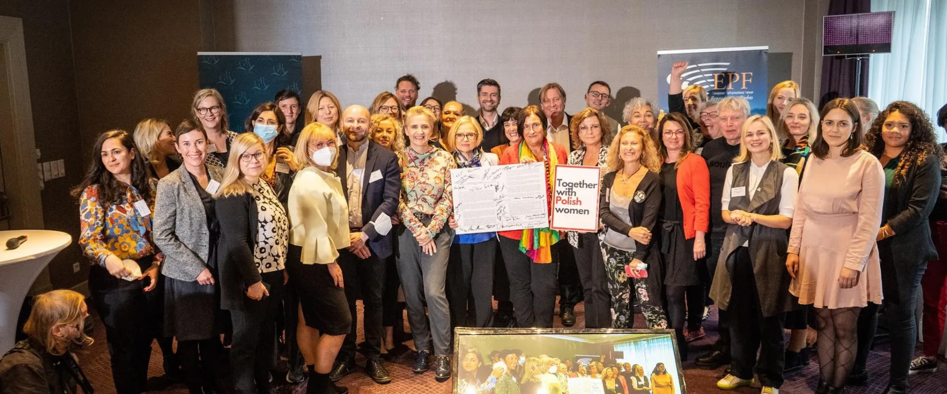 Duża grupa osób stojących razem w sali konferencyjnej podczas wydarzenia manifestu Warszawskiego, na środku trzyma podpisany dokument i plakat „Together with Polish women”, w tle widoczne banery organizacyjne.