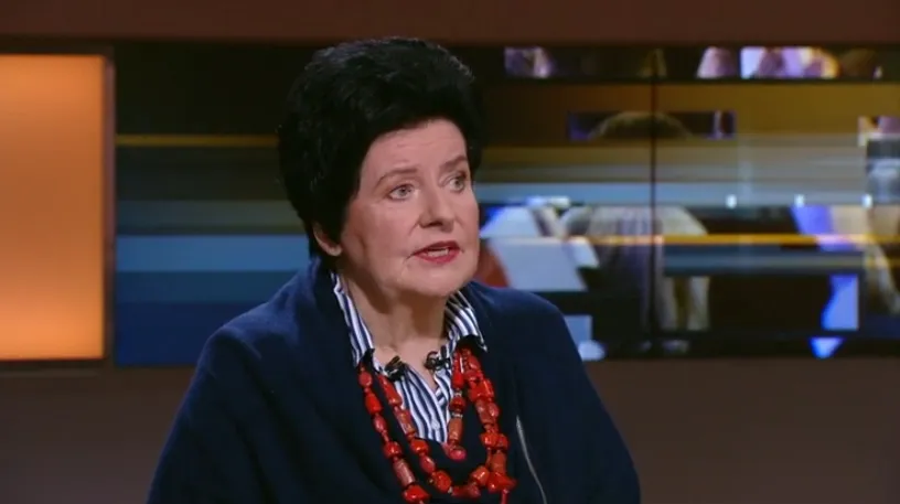 Kobieta w ciemnym garniturze i czerwonej biżuterii siedzi w studiu telewizyjnym i wygłasza komentarz.