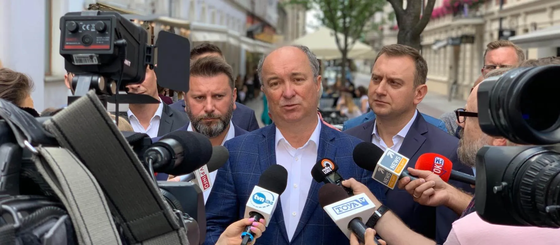 Polityk w granatowym garniturze udziela wywiadu przed kamerami, otoczony dziennikarzami i mikrofonami na ulicy.