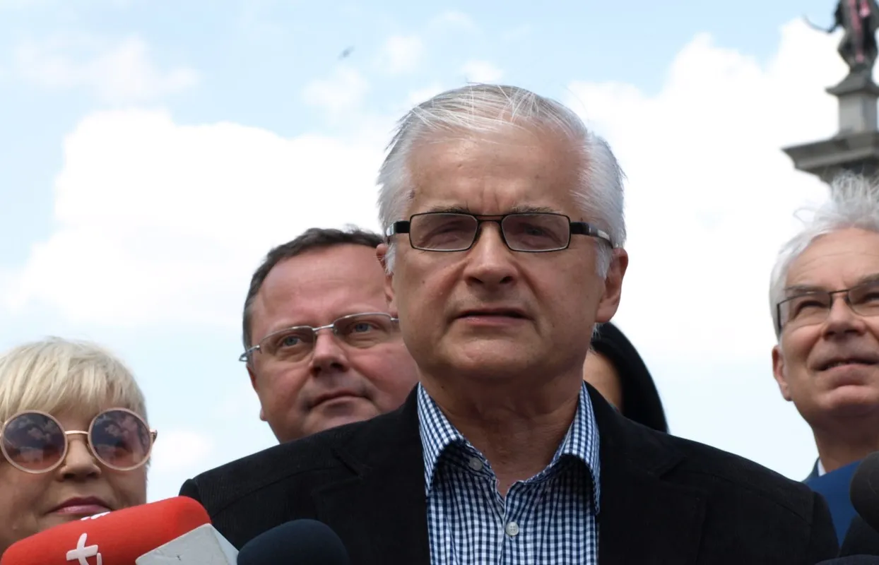 Mężczyzna z siwymi włosami i okularami przemawia na konferencji prasowej, stojąc przed mikrofonami, w towarzystwie innych osób na tle błękitnego nieba.