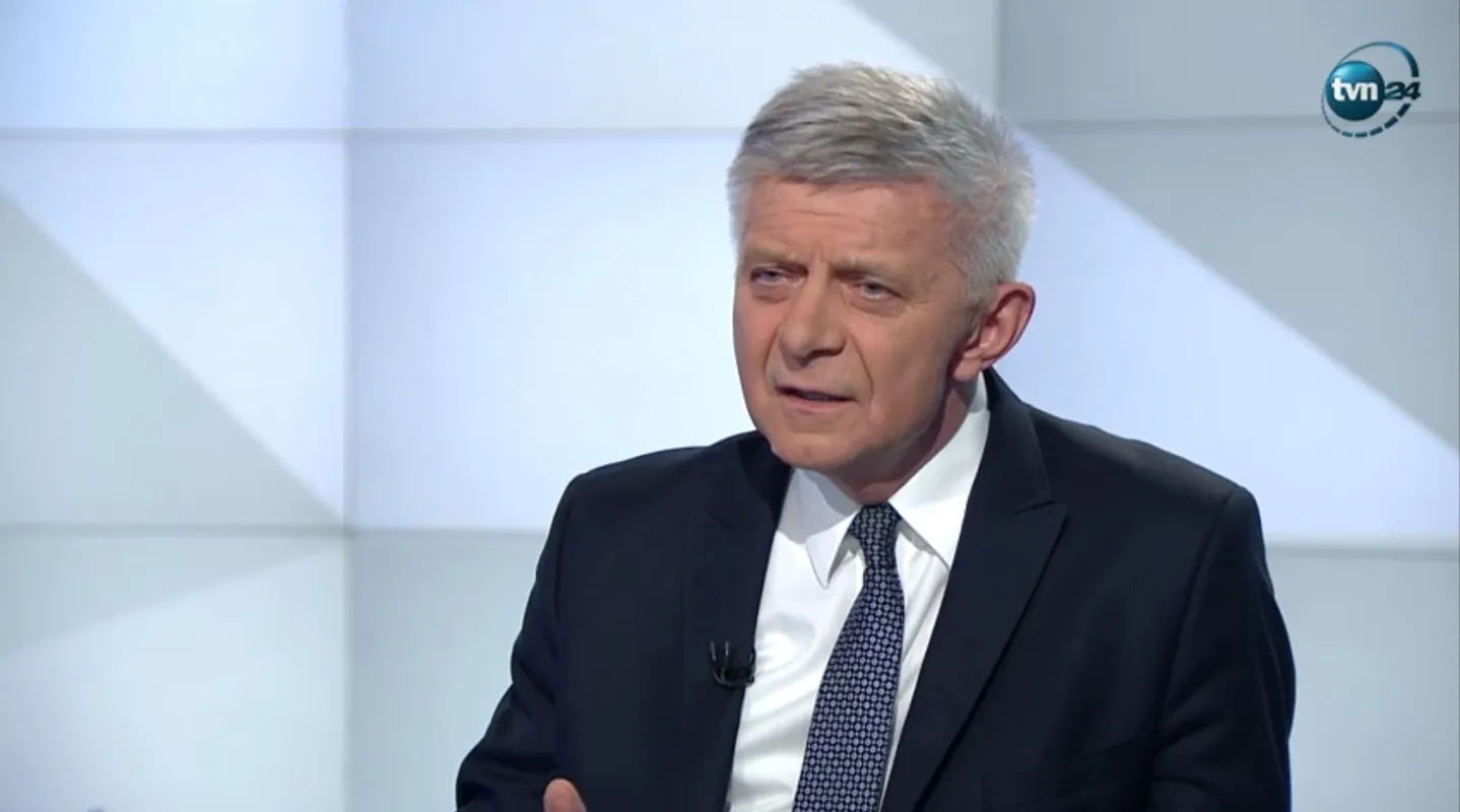 Poważny mężczyzna w garniturze w studiu telewizyjnym TVN24 wygłasza komentarz podczas programu.
