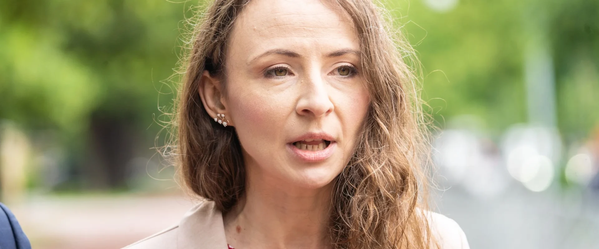 Kobieta z długimi, falującymi włosami mówi na zewnątrz, ubrana w beżowy żakiet i czerwono-białą bluzkę w drobny wzór, w tle zielona przestrzeń parkowa.