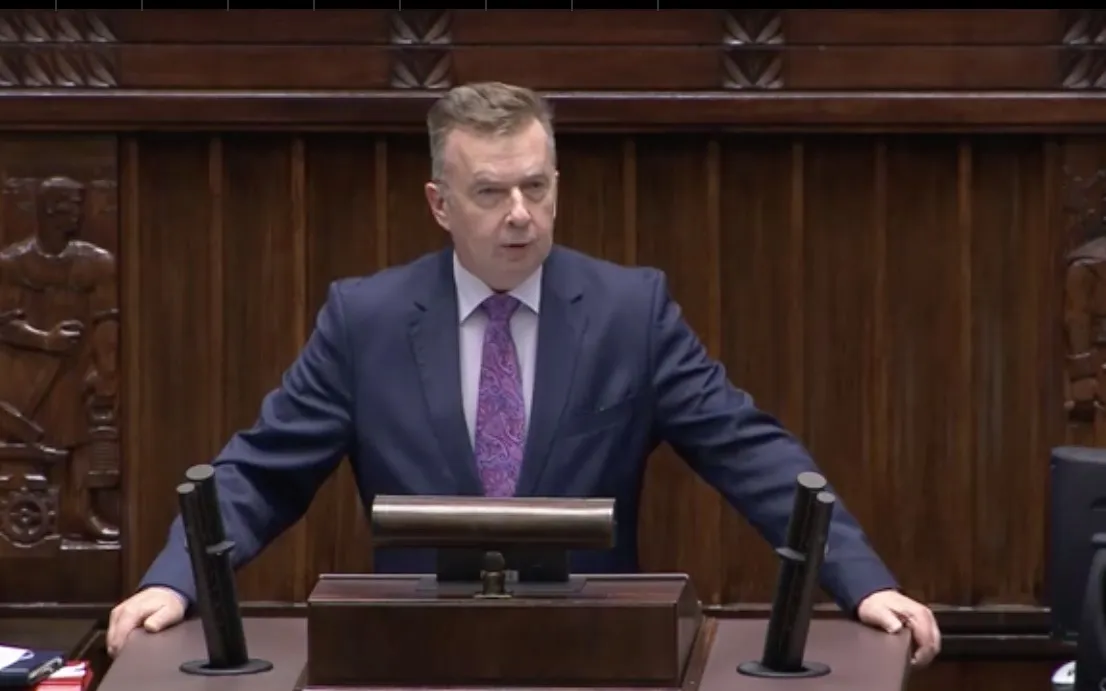 Polityk w granatowym garniturze i fioletowej krawacie przemawia zza mównicy w sali obrad.