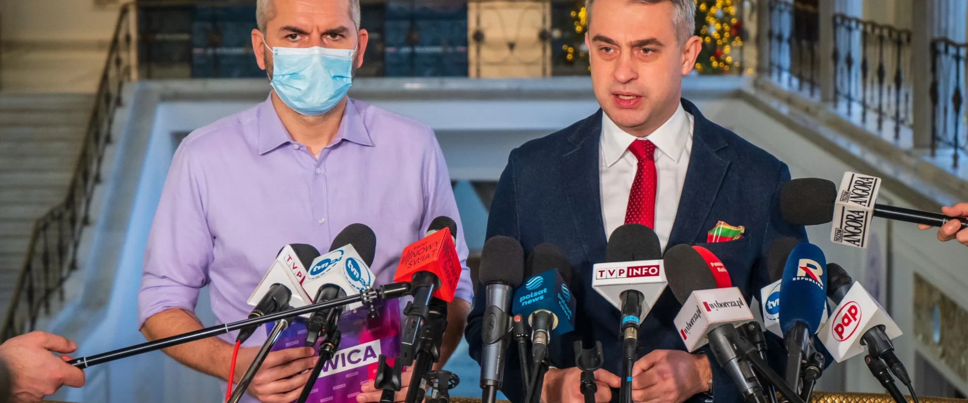 Dwóch mężczyzn w garniturach stoi za szeregiem mikrofonów podczas konferencji prasowej w eleganckim holu z choinką w tle; z lewej – w fioletowej koszuli i maseczce, z prawej – w granatowym garniturze z czerwonym krawatem.