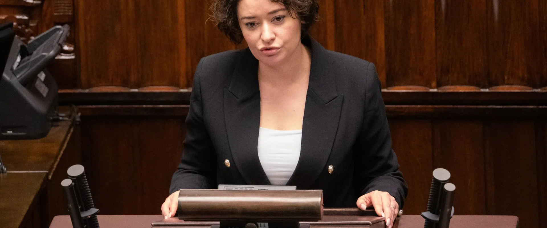 Kobieta z krótkimi kręconymi włosami stoi za mównicą w sali plenarnej, ubrana w czarny żakiet i białą bluzkę, wygłasza przemówienie.