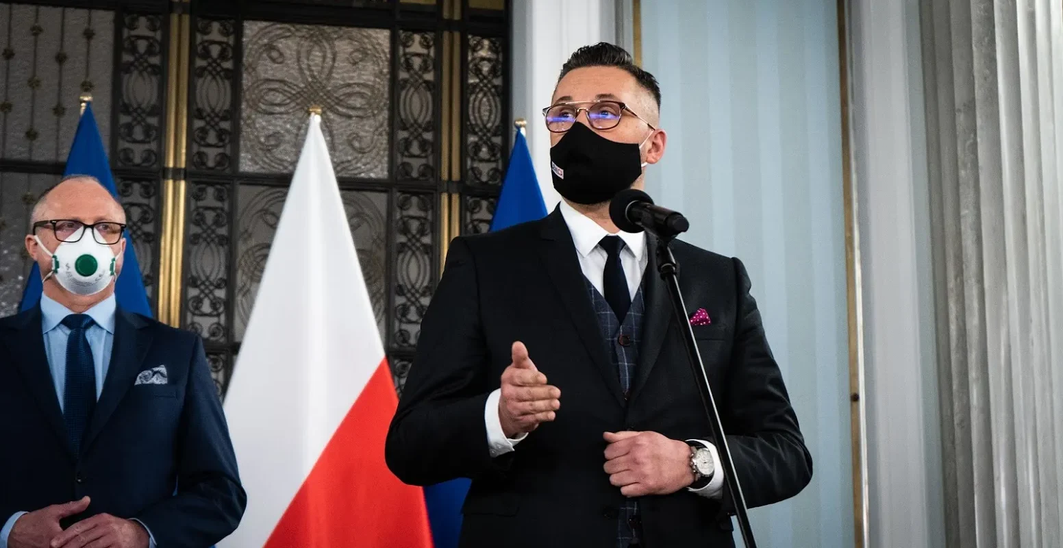 Mężczyzna w czarnym garniturze i maseczce przemawia do mikrofonu, stojąc obok drugiego mężczyzny w garniturze; w tle widoczne flagi Polski i Unii podczas wydarzenia „Wspólnie dla Polski”.