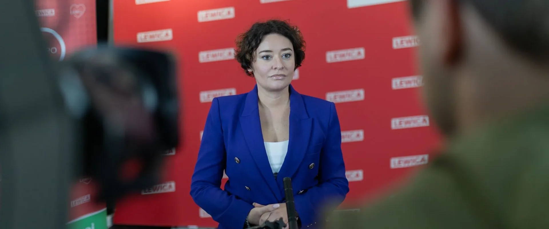 Kobieta w niebieskim garniturze przemawia podczas konferencji prasowej, stojąca przed czerwonym tłem z logo Lewica, otoczona kamerami.