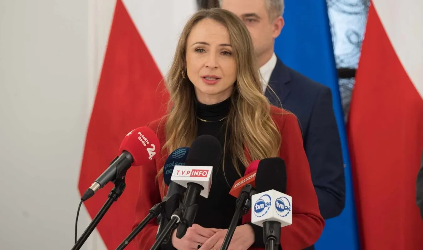 Kobieta w czerwonym płaszczu przemawia do mikrofonów podczas konferencji prasowej dotyczącej pomocy finansowej dla poszkodowanych przez powódź.