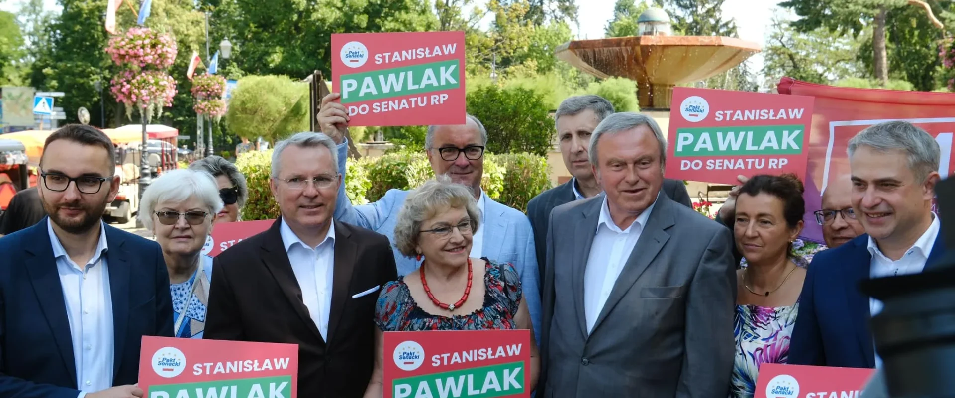 Grupa osób na plenerowej konferencji wyborczej stoi przed fontanną i trzyma plakaty z napisem Stanisław Pawlak do Senatu RP.