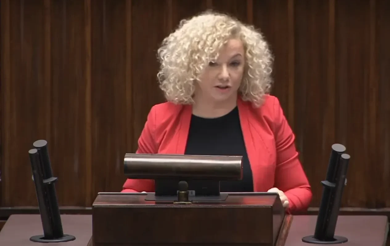 Kobieta o kręconych blond włosach w czerwonej marynarce stoi za podium i przemawia do publiczności.