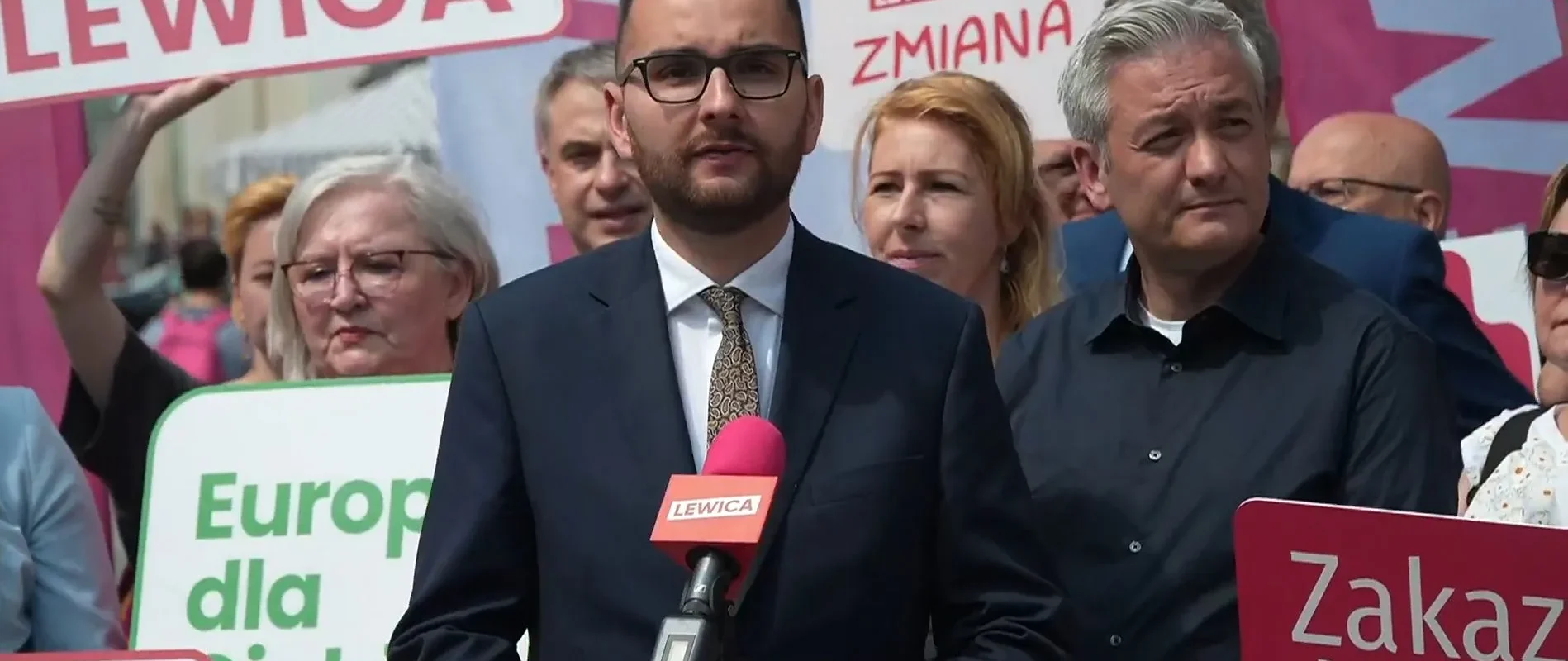 Polityk Kowal w garniturze przemawia na wiecu Lewicy, przedstawiając propozycję powołania Europejskiej Unii Energetycznej; w tle widoczni demonstranci i kolorowe banery.
