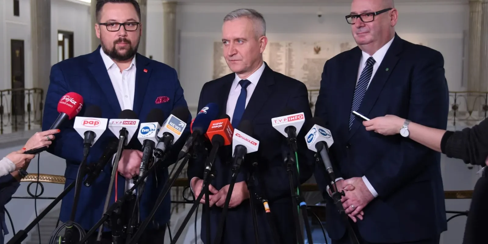Trzech mężczyzn w garniturach stoi przy licznych mikrofonach podczas konferencji prasowej w eleganckim wnętrzu z kolumnami w tle.