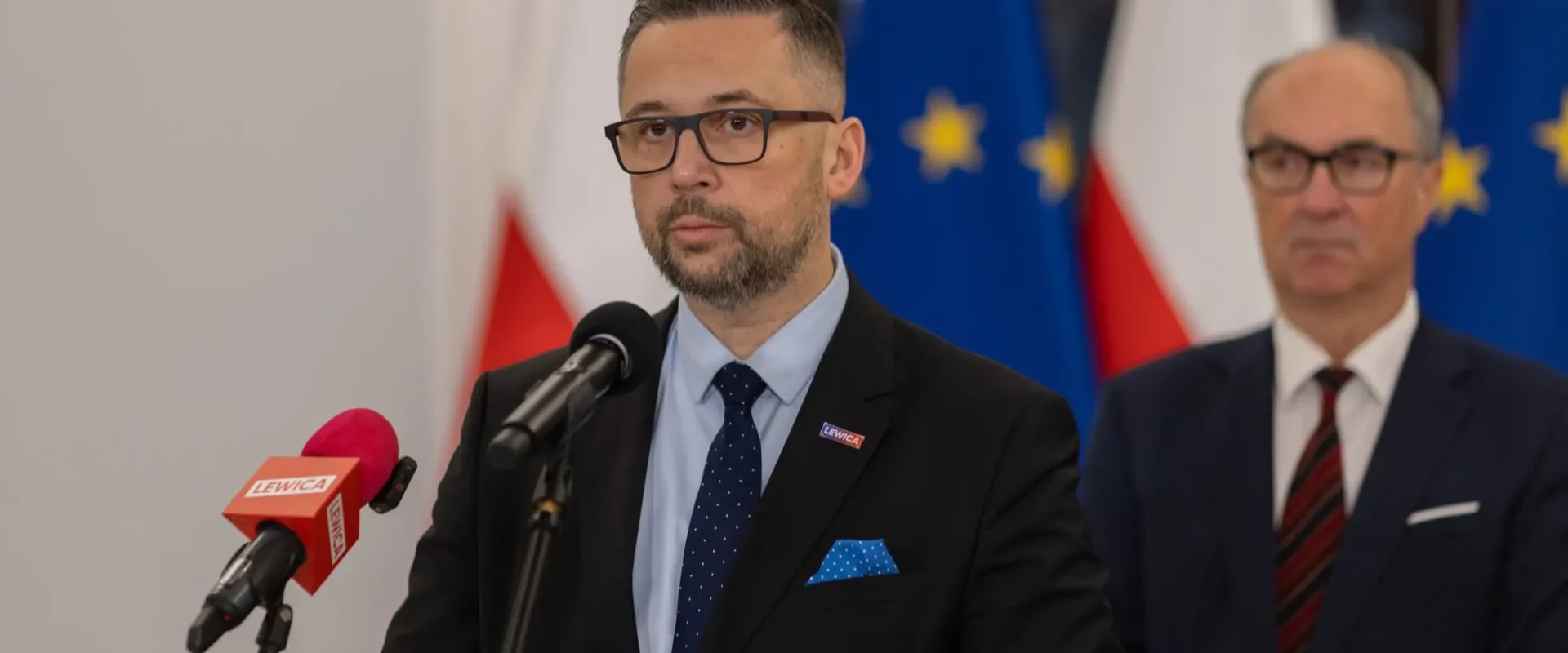 Mężczyzna w garniturze i okularach stoi przy mikrofonach podczas konferencji prasowej, z flagami państwowymi w tle (minister nauki i szkolnictwa wyższego).